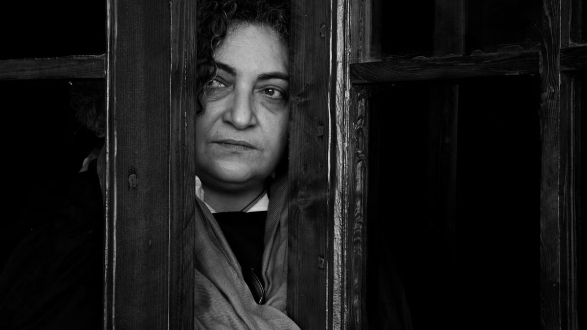 Black and white portrait of an elderly woman peering through a vintage wooden doorway.